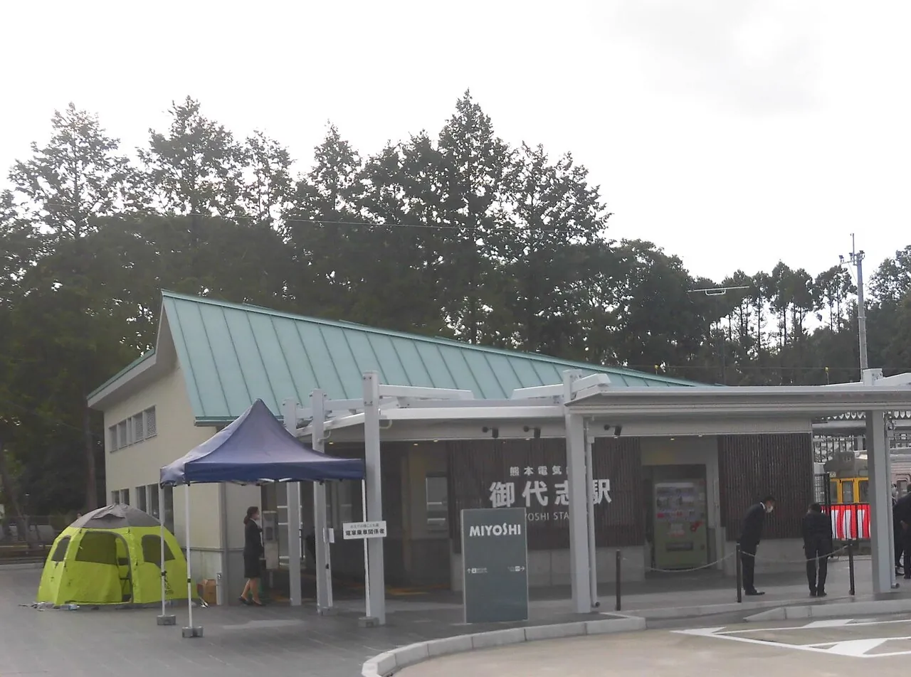 緑色の屋根の御代志駅の駅舎。駅名の看板と、木立が見える