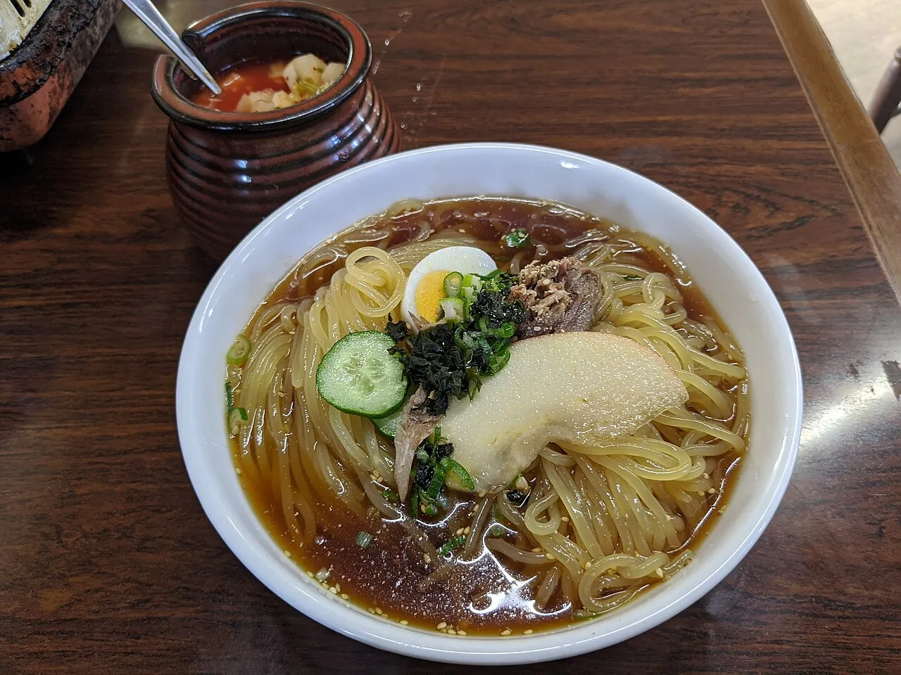 白い丼に盛られた冷麺。ゆで卵、きゅうり、のり、果物のスライスがのり、丼の奥に赤いスープの入った壺が見える