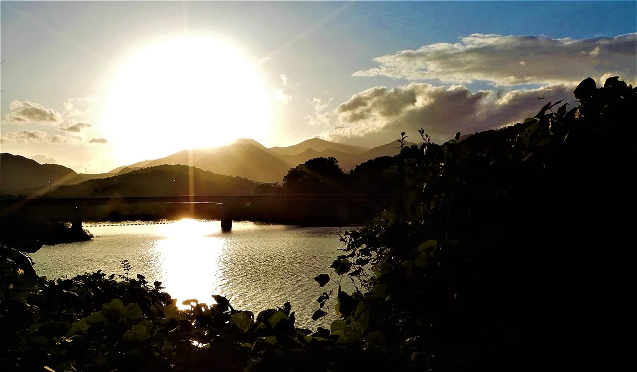 夕日が山の向こうに沈み、湖の水面に光が映る夕方の景色。手前に橋がかかる
